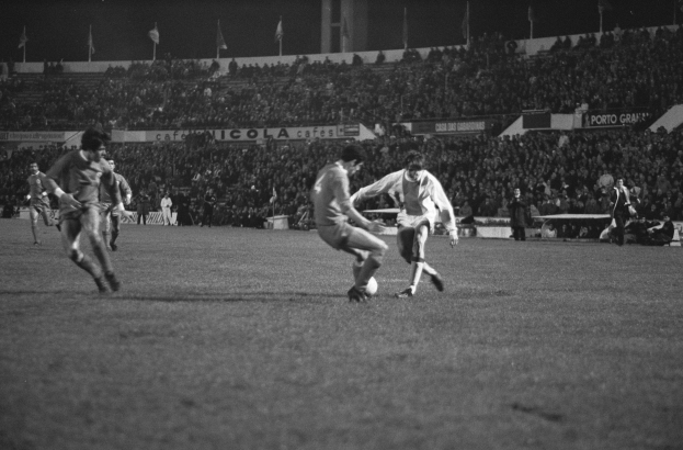 Schwarzes und weißes Bild von Männern, die Fußball auf einem Feld mit Zuschauern auf den Rängen und Fahnen im Hintergrund spielen.