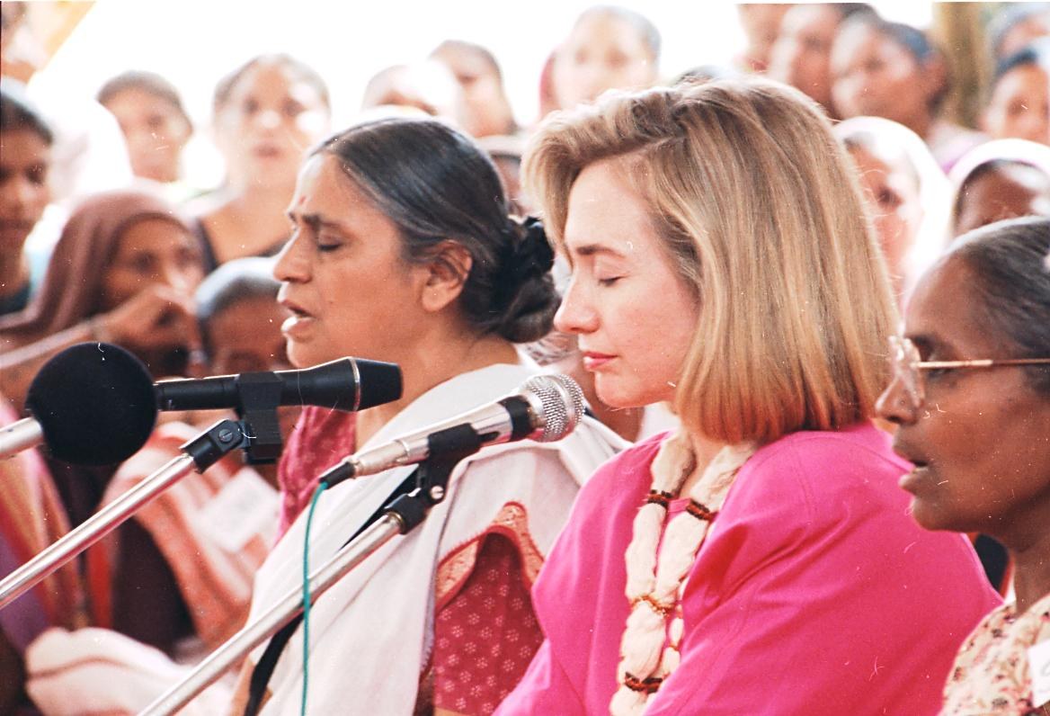 Zwei Frauen vor einem Publikum mit Mikrofonen, eine spricht, während die andere zuhört.