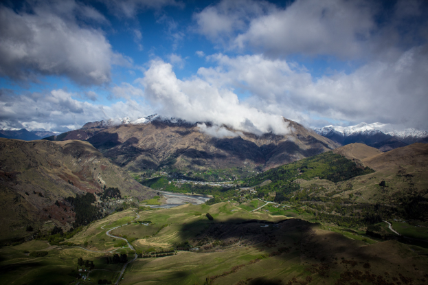 Luftaufnahme von Queenstown, Neuseeland, mit grünem Gras, Bäumen, einer gewundenen Straße und weißen Wolken am Himmel.