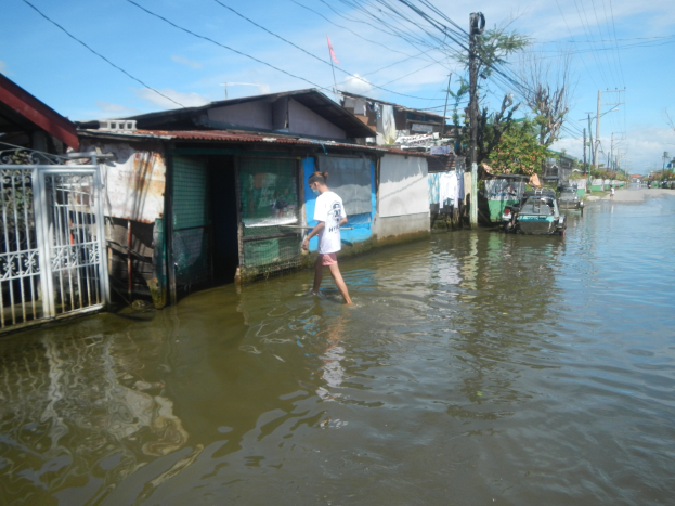 Ein Mann in einem weißen T-Shirt und Shorts watet durch eine überflutete Straße in Manila, Philippinen, umgeben von Häusern, Bäumen, Pfählen, Drähten und Fahrzeugen unter einem sichtbaren Himmel.