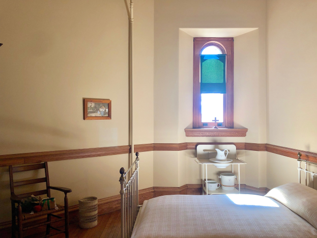 Ein Schlafzimmer mit einem Bett, einem Stuhl, einem Korb, einer Stange, einem Foto Rahmen, einem Fenster, einem Tisch mit einer Kanne und Tassen und einem Dach.