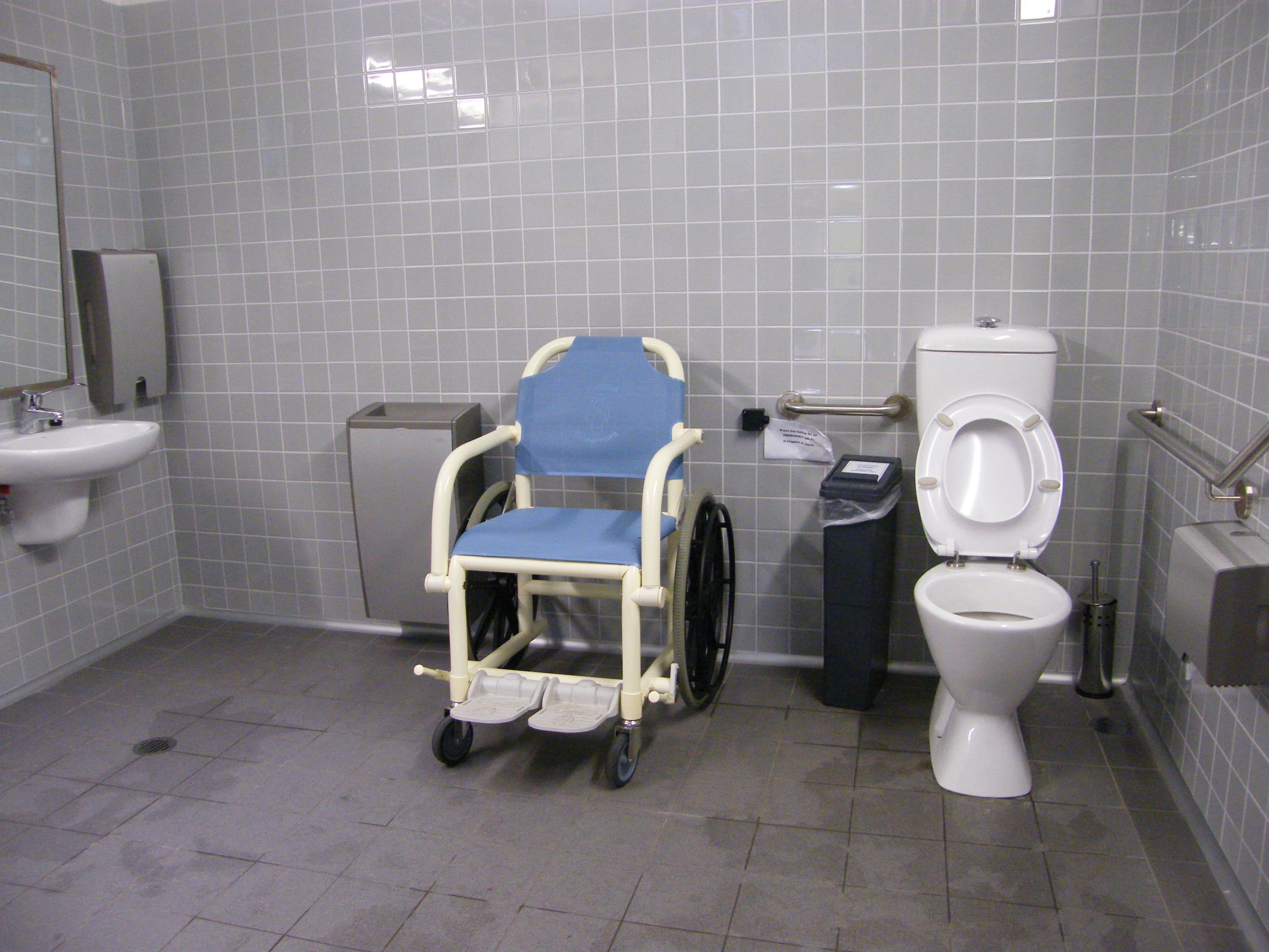 Ein Badezimmer mit Toilette, Kommode, Spülkasten, Mülltonne, Duschbrause, Spiegel, Waschbecken und anderen Gegenständen; die Wände sind gefliest, der Boden ist unten sichtbar und ein Rollstuhl steht neben der Kommode in der Mitte des Raumes.