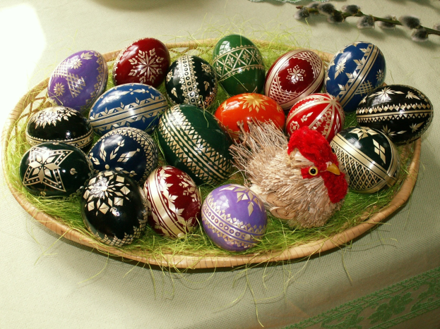 A woven basket containing decorated Easter eggs sits on a table draped with a cloth, with a toy in the basket's center and flowers to the right.
