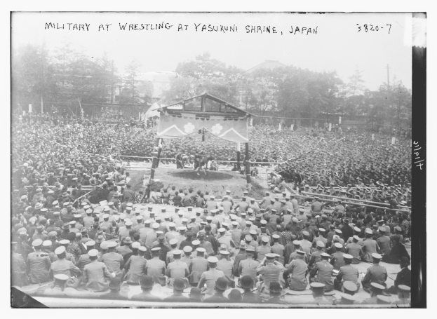 Schwarzes und weißes Foto einer Menge, die einen Wrestling-Kampf auf einer Bühne im Yasukuni-Schrein, Japan, beobachtet, mit Bäumen und Gebäuden im Hintergrund und Text oben und unten.
