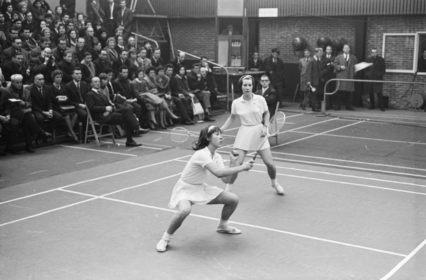 Two women playing tennis on a court surrounded by a fence, with spectators seated and standing in the background, and a building visible behind the fence; the image is in black and white.