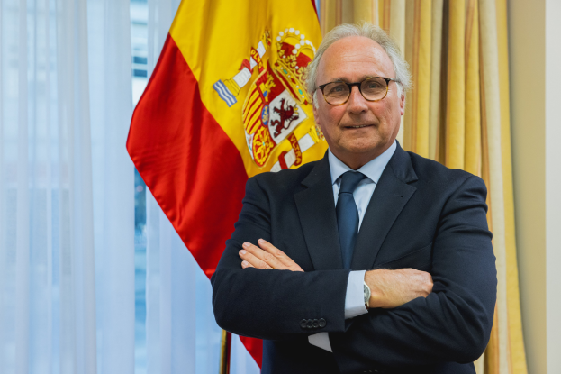 Ein Mann im Anzug und Krawatte, der eine Brille und eine Uhr trägt, steht lächelnd vor einer spanischen Flagge in einem formellen Setting mit Vorhängen und einer Wand im Hintergrund.