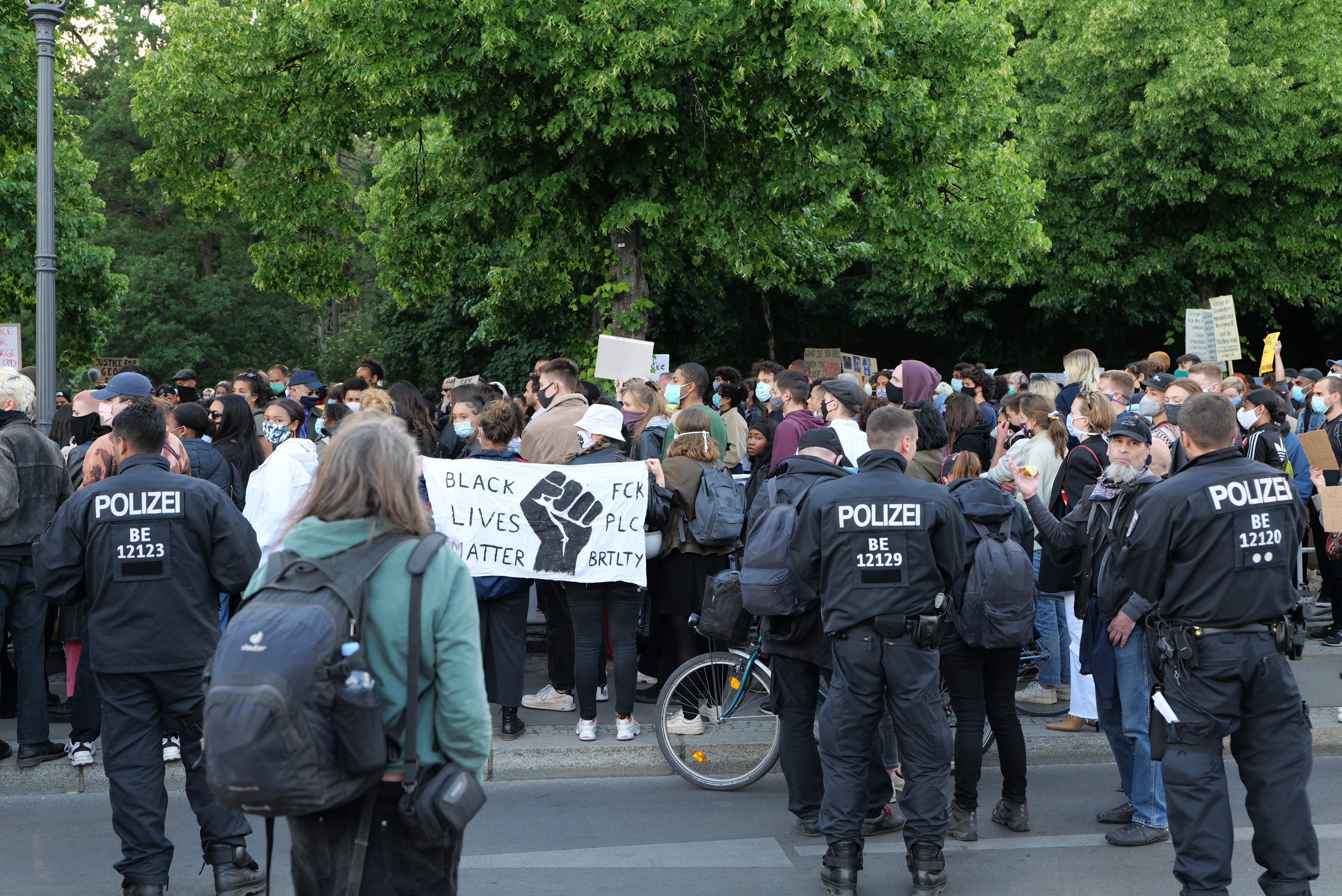 Eine große Gruppe von Menschen steht an der Seite einer Straße, einige halten Schilder, mit einem Fahrrad im Vordergrund und Bäumen und einem Pfahl im Hintergrund, an einer Black Lives Matter Demonstration in Berlin teilnehmend.