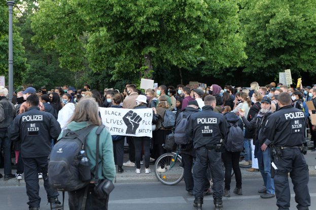 Eine große Gruppe von Menschen steht an der Seite einer Straße, einige halten Schilder, mit einem Fahrrad im Vordergrund und Bäumen und einem Pfahl im Hintergrund, an einer Black Lives Matter Demonstration in Berlin teilnehmend.