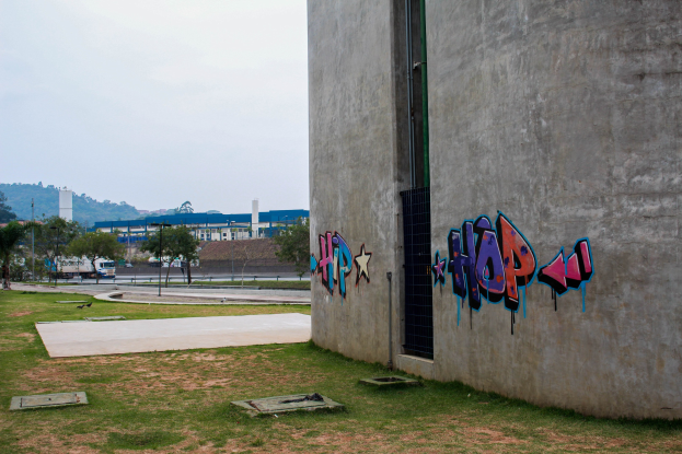 Betongebäude mit Graffiti an der Seite, umgeben von Gras, Bäumen, Strommasten, Fahrzeugen auf der Straße, anderen Gebäuden, fernen Hügeln und einem klaren blauen Himmel.
