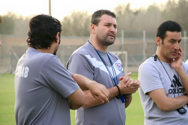 Eine Gruppe von M├Ąnnern in grauen T-Shirts auf einem Fussballfeld mit einem unscharfen Hintergrund aus Netz, Pfosten und B├Ąumen, einer Person als Trainer identifiziert.