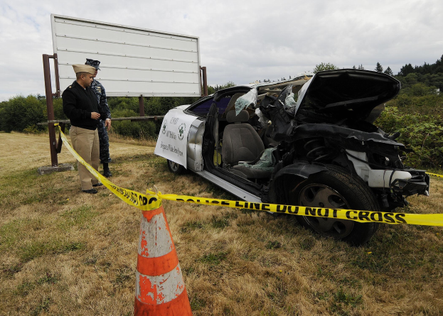 Polizist in Uniform inspiziert ein schwer beschädigtes Auto mit eingedrückter Motorhaube und zersplitterten Fenstern am Straßenrand, mit einem Verkehrskegel und einer Schnur in der Nähe vor einem Hintergrund aus Bäumen und einem bewölkten Himmel.