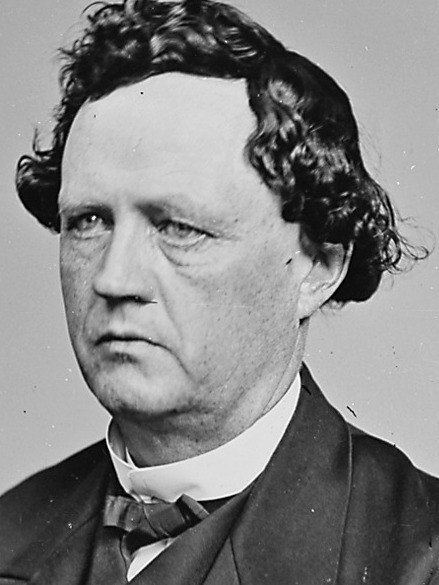 Schwarzes und weißes Foto eines Mannes mit lockigen Haaren in einem Anzug, der formell mit Händen in den Taschen und einem ernsten Gesichtsausdruck steht.