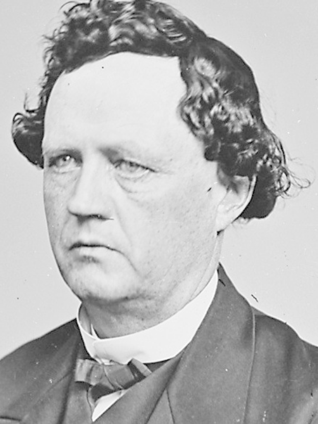 Schwarzes und weißes Foto eines Mannes mit lockigen Haaren in einem Anzug, der formell mit Händen in den Taschen und einem ernsten Gesichtsausdruck steht.