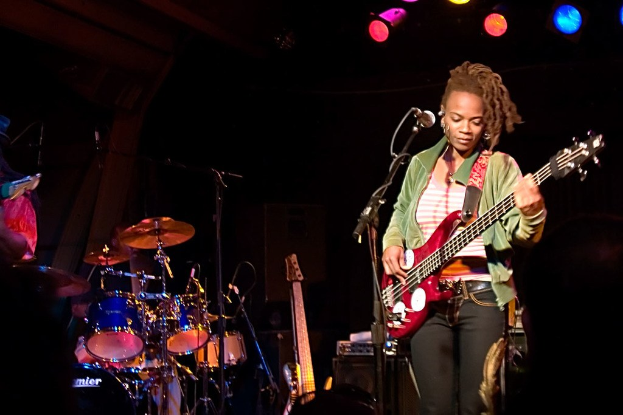 Eine Frau in einer gr├╝nen Jacke spielt Gitarre auf der B├╝hne w├Ąhrend eines Konzerts, mit einer Bandaufstellung zu ihrer Linken und Lichtern ├╝ber einem dunklen Hintergrund.