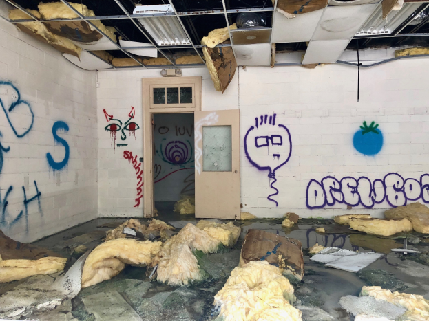 Ein Raum mit Graffiti an den Wänden, eine Tür im Hintergrund, Deckenstangen und Lampen sowie verschiedene Gegenstände auf dem Boden verteilt.