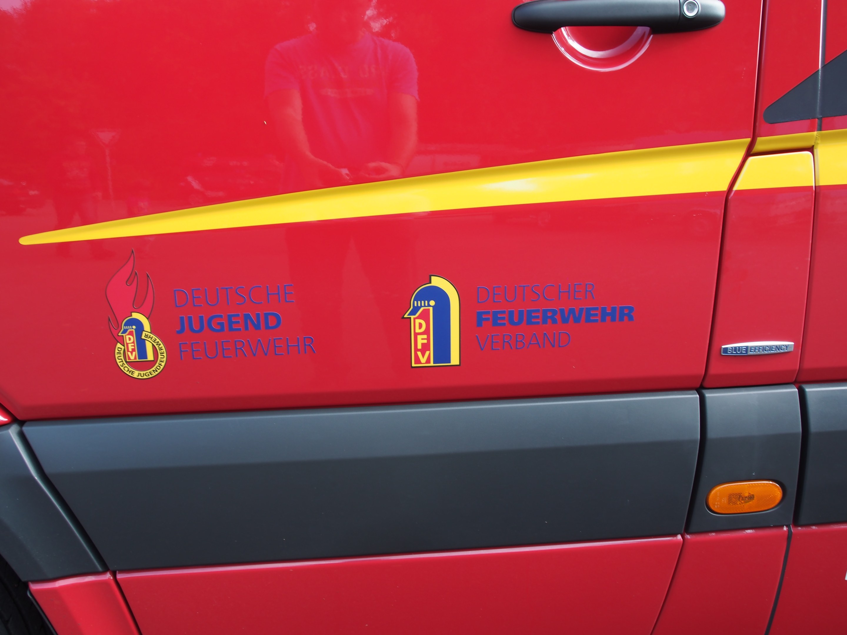 Red van with a yellow stripe, door handle, and a shield logo featuring a lion in a laurel wreath, labeled "Deutsche Jugend Feuerwehr Verband".
