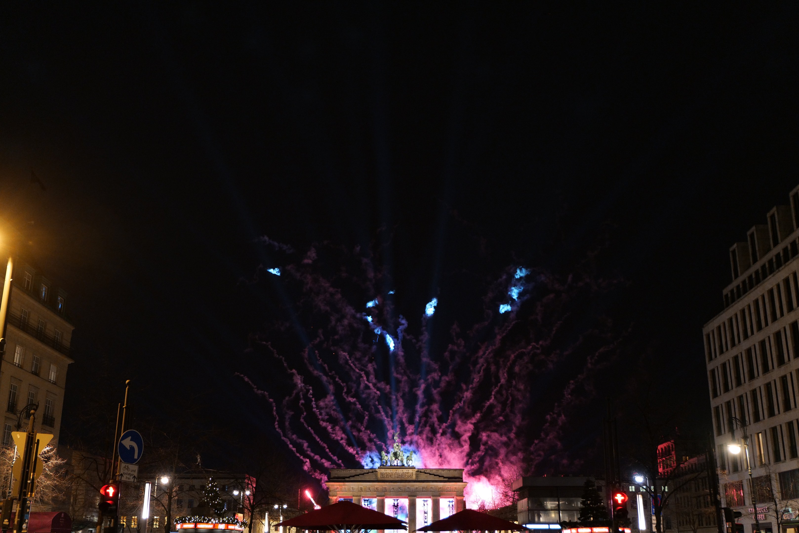 Eine belebte Stadtstraße in Berlin, Deutschland, mit beleuchteten Gebäuden, Pfählen, Schildern, Verkehrszeichen, Zelten und Fahrzeugen, mit dem Brandenburger Tor im Hintergrund und Feuerwerk, das den Nachthimmel erhellt.