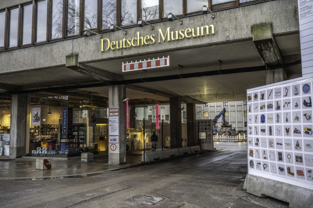 Außenansicht des Deutschen Museums in Berlin mit einem großen Gebäude mit Glasfenstern, Säulen und einer Namensschilder, sowie einer Informationsanzeige und umliegenden städtischen Elementen.