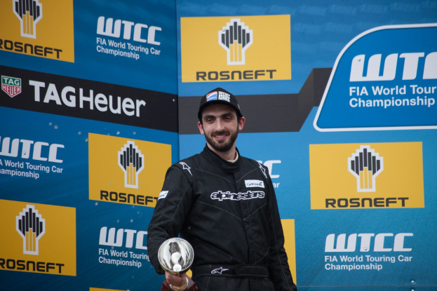 Ein Mann in schwarzer Kleidung und einer Mütze steht auf einem Podium, lächelt und hält eine Trophäe in der Hand, während im Hintergrund eine blaue Tafel mit der Aufschrift "FIA World Touring Car Championship" zu sehen ist.