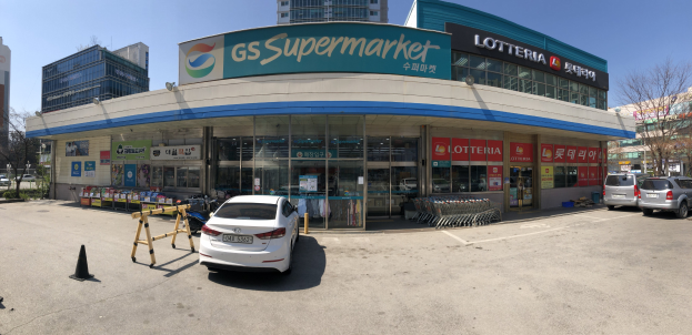 Ein Auto steht vor einem Geschäft mit der Aufschrift "GS Supermarkt" zusammen mit anderen Fahrzeugen, Verkehrskegeln und verstreuten Gegenständen in der Nähe, vor einem Hintergrund aus Gebäuden, Bäumen und einem klaren blauen Himmel.