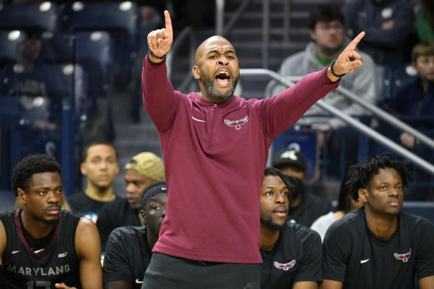 Cheftrainer der Maryland-Männer-Basketballmannschaft in einem rotbraunen Hemd und schwarzen Shorts in einer Diskussion auf der Seitenlinie eines Basketballplatzes umgeben von sitzenden und stehenden Personen.