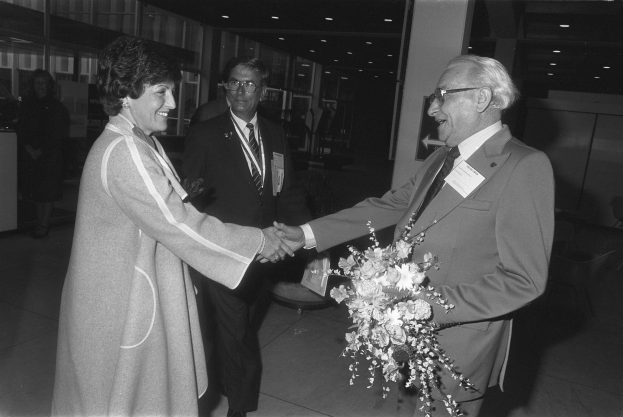 Ein Mann im Anzug und mit Krawatte gibt einer lächelnden Frau in einem Flughafen die Hand, die einen Blumenstrauß hält, während im Hintergrund andere Personen zu sehen sind.