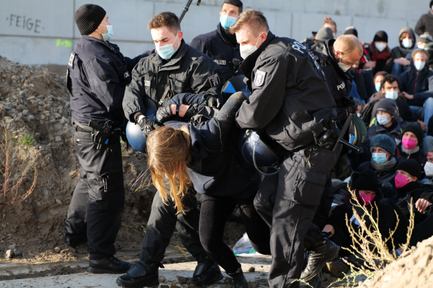 Gruppe maskierter Individuen in Kapuzenpullis, einige bewaffnet, umgeben eine Frau, die von der Polizei eskortiert wird, mit städtischer Vegetation und einer Wand mit Protesttext im Hintergrund.