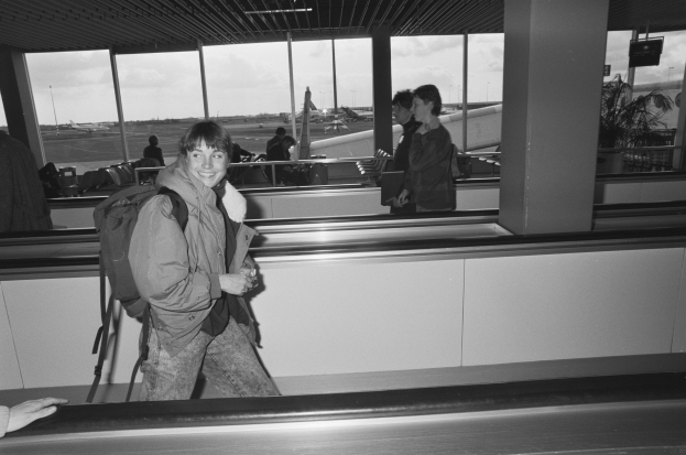 Eine Frau, die lachend durch einen Flughafen geht und von Menschen, Flugzeugen, Pfählen, Bäumen und einem klaren blauen Himmel umgeben ist.