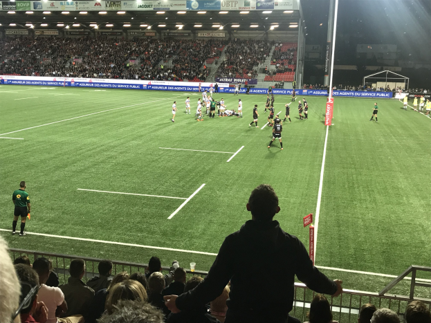 Eine Gruppe von Menschen, die Rugby in einem vollen Stadion mit saftig grünem Rasen spielen, umgeben von Texttafeln und beleuchtet von Stadionlampen, mit einem zentralen Pfosten auf dem Feld.