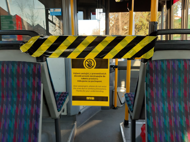 Innenansicht eines Busses mit gelb-schwarzem Absperrband, Sitzen, Stangen, Glasfenstern und sichtbaren Bäumen, Gebäuden und Himmel draußen.