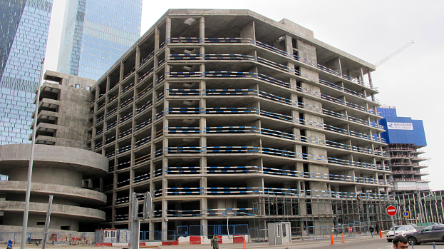 Ein großes Bauwerk im Bau in einer städtischen Umgebung, umgeben von Wolkenkratzern, mit Fahrzeugen, Fußgängern, Verkehrsinfrastruktur und Kränen unter einem klaren Himmel.