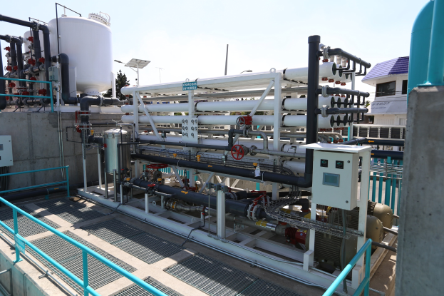 Large industrial water treatment plant with extensive piping, valves, machinery, railings, buildings, trees, and a clear blue sky in the background.