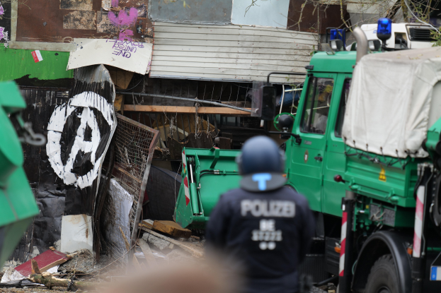Ein Polizist steht vor einem Gebäude mit Graffiti, umgeben von Fahrzeugen, Bäumen und anderen Gegenständen; ein menschliches Gesicht ist unten teilweise sichtbar.