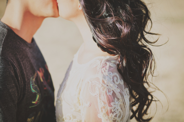 A couple in wedding attire embraces and kisses during sunset, with the woman in a white dress and the man in a black t-shirt, set against a warm, blurred background.