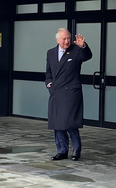 Prinz Philip, Herzog von Edinburgh, in einem Anzug und Krawatte, winkt einer Menge zu, während er in einem Gebäude mit einer Tür und einer Texttafel im Hintergrund steht.