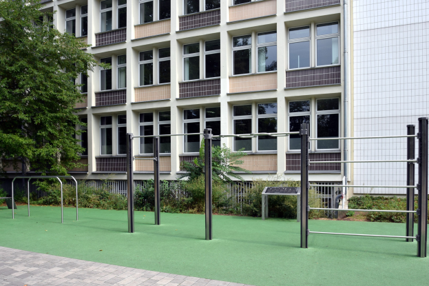 A multi-story building with balconies and railings, surrounded by greenery, featuring an artificial turf playground in the foreground.