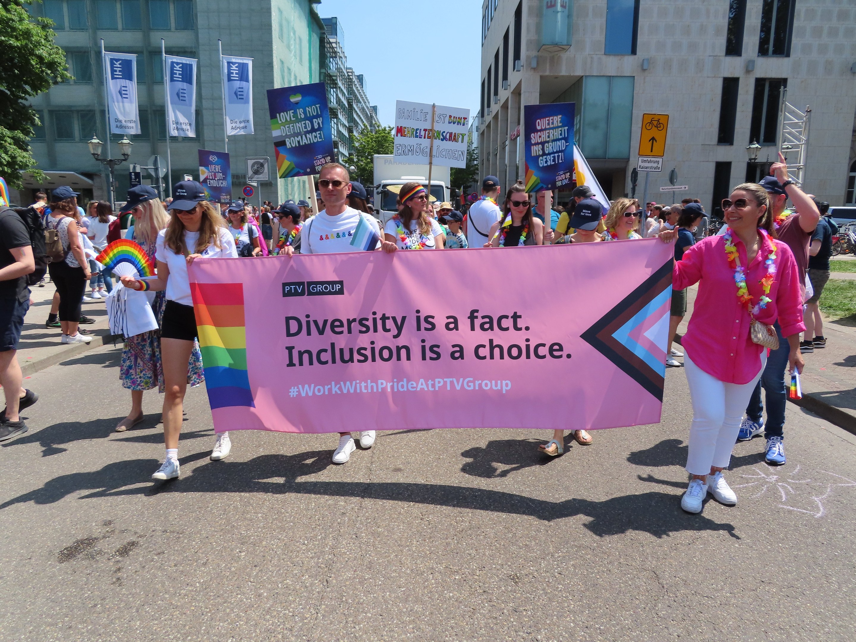 Eine bunte Gruppe von Menschen marschiert auf einer Straße, hält ein Banner mit der Aufschrift "Diversität ist eine Tatsache, Inklusion ist eine Wahl", einige tragen Mützen und Schutzbrillen, andere zusätzliche Banner, vor einem Hintergrund aus Gebäuden, Bäumen, Laternenpfählen und einem klaren blauen Himmel.