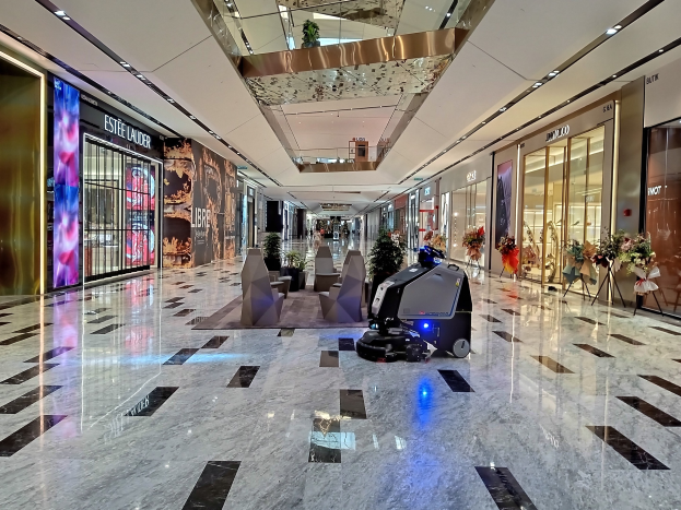 A robot vacuum cleaner moves across a shopping mall floor, surrounded by decorative plants, seating, floral arrangements, glass doors, informational boards, and overhead lighting.
