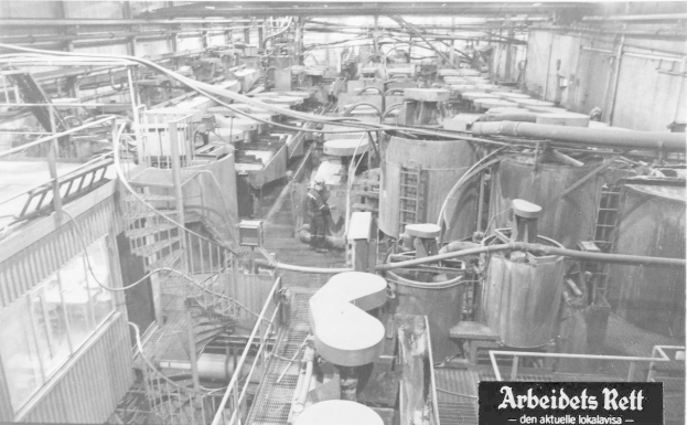 Schwarzes und weißes Foto eines großen industriellen Fabrikinneren mit einer Person zwischen Maschinen, Rohren und baulichen Elementen, mit dem Text "Arbeidets Rett, die größte Fabrik der Welt" unten.