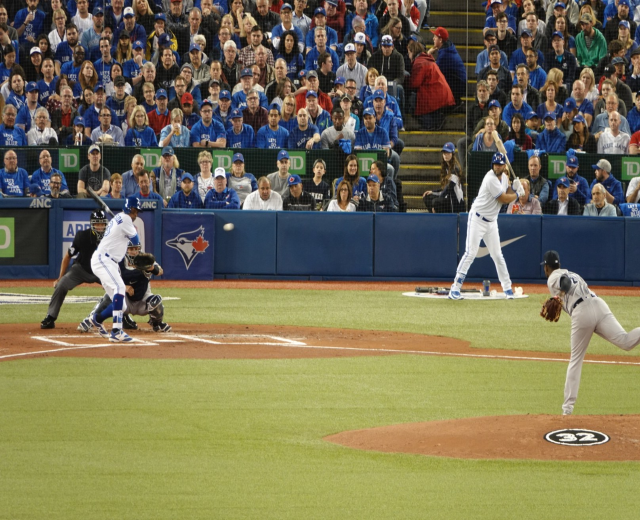 Baseballspieler schwingt einen Schläger auf einem Feld mit Teamkollegen in Sportuniformen und Zuschauern im Hintergrund.