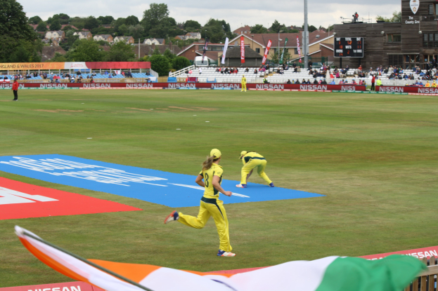 Cricket-Spieler im Einsatz während des ICC Women's T20 World Cup 2019-Spiels zwischen England und Australien, mit Zuschauern, Werbetafeln, Fahnen und einem Scoreboard im Hintergrund.