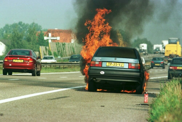 Ein Auto, das auf der Straße in Flammen steht, umgeben von anderen Fahrzeugen, umgeben von Bäumen, Gebäuden und einem klaren blauen Himmel, mit Gras und einem Feuerlöscher auf der rechten Seite.