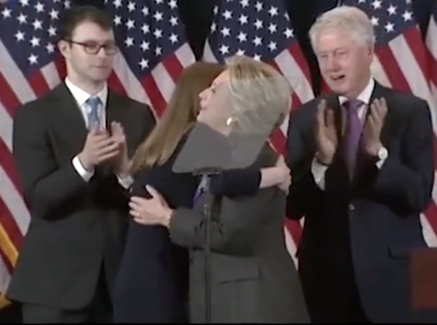 Hillary Clinton und Bill Clinton vor einem Rednerpult mit Mikrofon stehend, mit klatschenden Händen und mehreren Flaggen im Hintergrund.