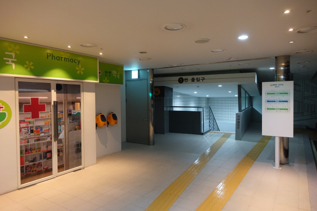 Apotheken-Eingang mit einem "Apotheke"-Schild auf der linken Seite, einer Tafel mit Text auf der rechten Seite, beleuchteten Lampen darüber und Hintergrundgegenständen.