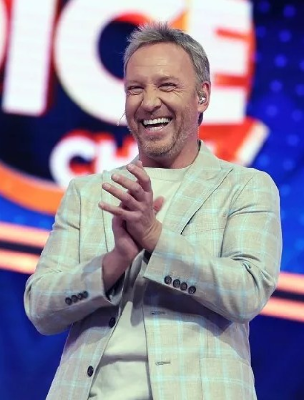 Ein Mann in einem Anzug auf einer Bühne, der mit einem breiten Lächeln klatscht, vor einer Tafel mit der Aufschrift "The Voice."