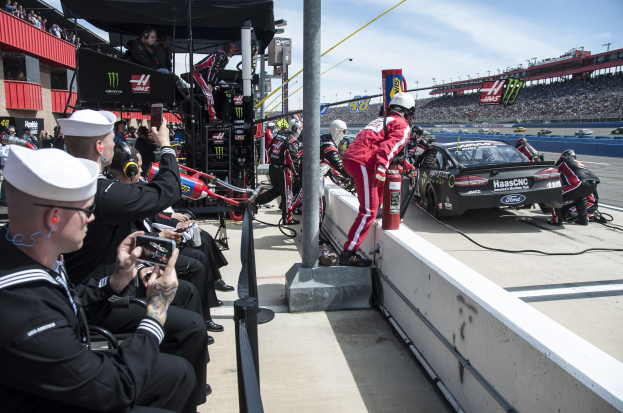 Gruppe von Menschen um ein Rennauto auf einer Rennstrecke, einige tragen Kappen und halten Kameras, mit einem Pfahl im Vordergrund und einem Gebäude mit Geländern im Hintergrund, Zuschauer auf Tribünen unter einem bewölkten Himmel.