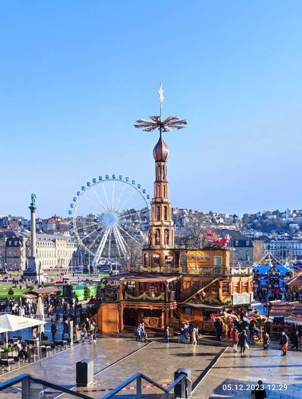 Ein belebter Stadtplatz mit einem zentralen Riesenrad, umgeben von Gebäuden, Bäumen und Menschen, unter einem klaren blauen Himmel, was auf eine Weihnachtsmarkt-Szenerie hindeutet.
