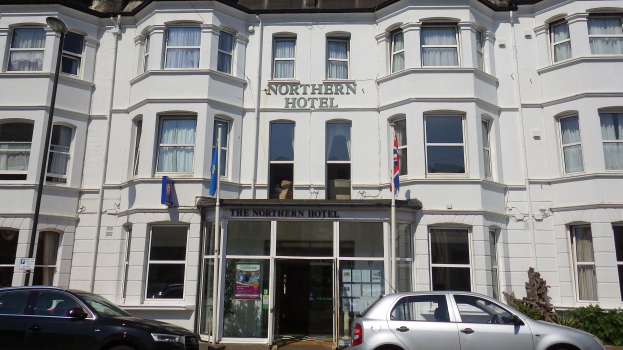 Das Northern Hotel in Brighton, England, ein großes weißes Gebäude mit Glasfenstern und -türen, das eine Schildertafel und zwei geparkte Autos vorweist, mit Straßenmerkmalen wie einem Pfahl, Pflanzen und Fahnen unter einem bewölkten Himmel.