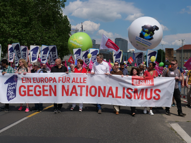 A group of people marching in a demonstration against nationalism in Berlin, carrying banners, flags, and balloons, with trees, buildings, and a cloudy sky in the background.