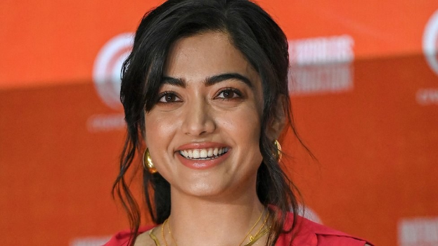 Eine Frau in einem roten Kleid mit einem eleganten Dutt und dezentem Make-up lächelt in die Kamera, trägt schwarze High Heels und hat ein hellgelbes Banner mit der Aufschrift "Sonam Kapoors schönste Looks vom Cannes Film Festival" im Hintergrund.